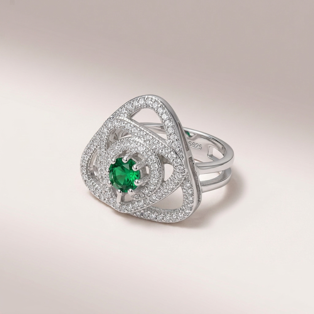 A silver-toned geometric ring featuring a central round green emerald stone surrounded by pavé-set clear stones, photographed on a soft light beige studio background.