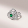 A silver-toned geometric ring featuring a central round green emerald stone surrounded by pavé-set clear stones, photographed on a soft light beige studio background.
