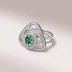 A silver-toned geometric ring featuring a central round green emerald stone surrounded by pavé-set clear stones, photographed on a soft light beige studio background.